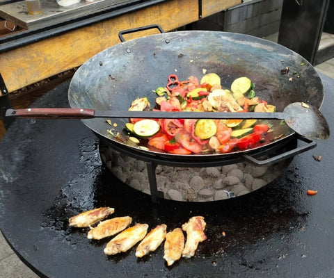 Eingebrannte Fyreza Wok-Pfanne 55 cm mit Kartoffeln direkt auf OFYR Feuerplatte – ideal für Kochen über offenem Feuer