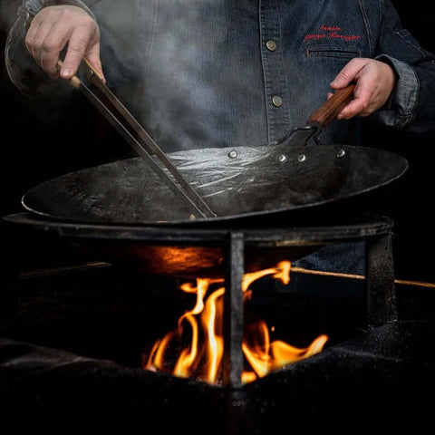 Ein Koch, der eine handgeschmiedete Wok-Pfanne über einem offenen Feuer verwendet, um köstliche Gerichte zuzubereiten.